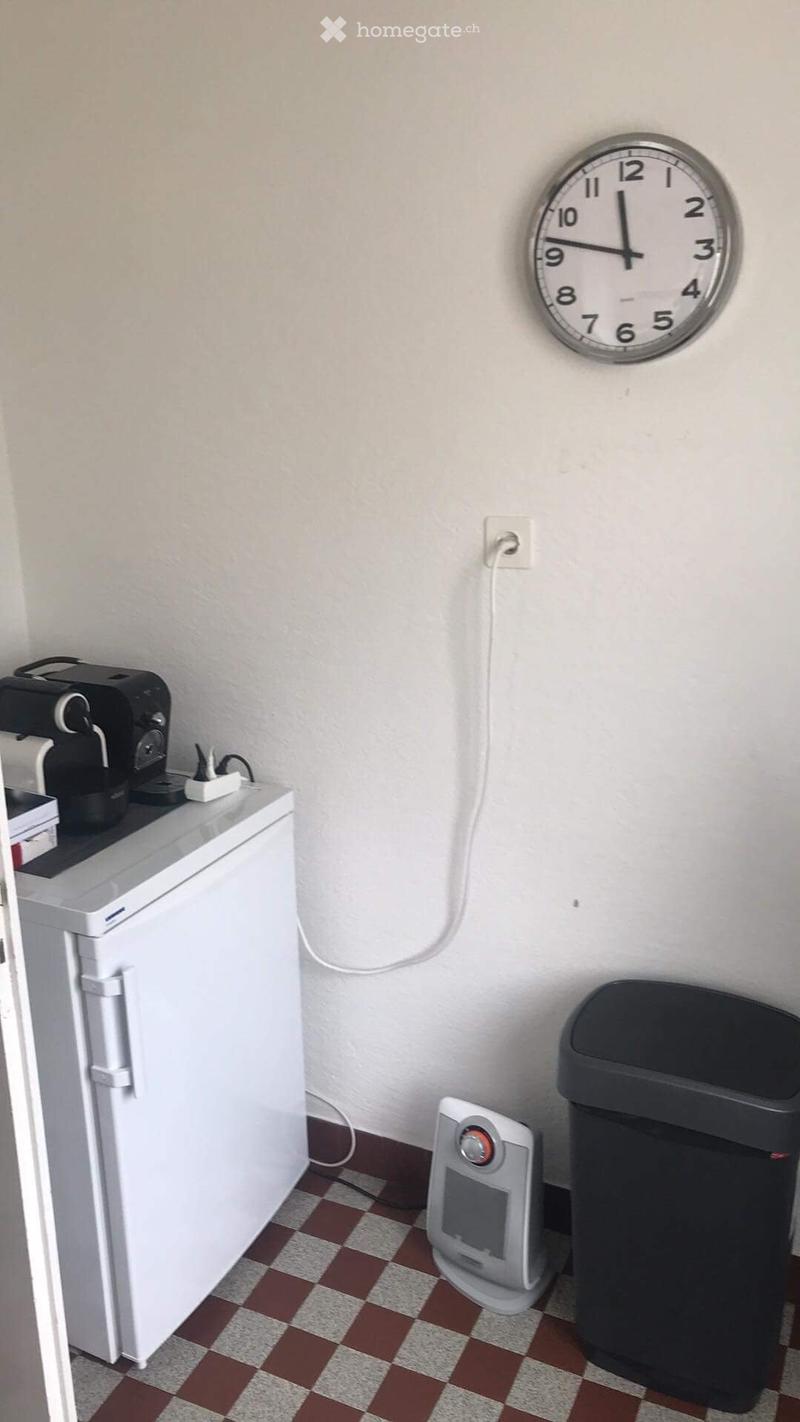 A small kitchen with a white refrigerator, a coffee maker on top, a heater, a trash can, and a wall clock.