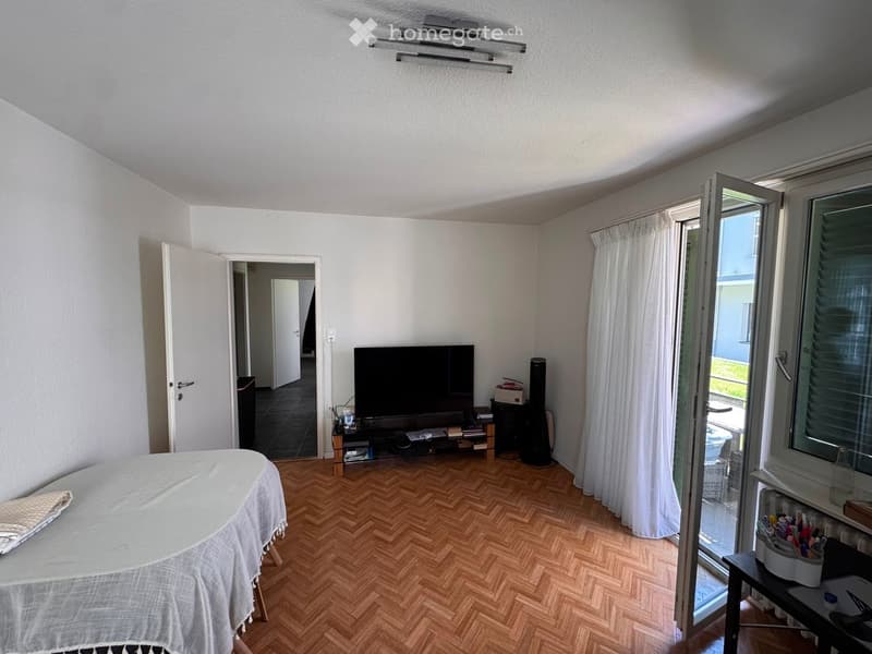 A furnished room with a wooden herringbone floor, a covered table, a television on a stand, and a large window with white curtains.