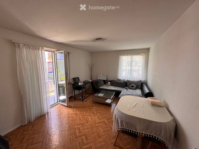 A cozy living room with a sectional sofa, coffee table, desk, and a small table covered with a cloth. The room features wooden flooring, large windows with curtains, and a sliding glass door leading to a balcony.