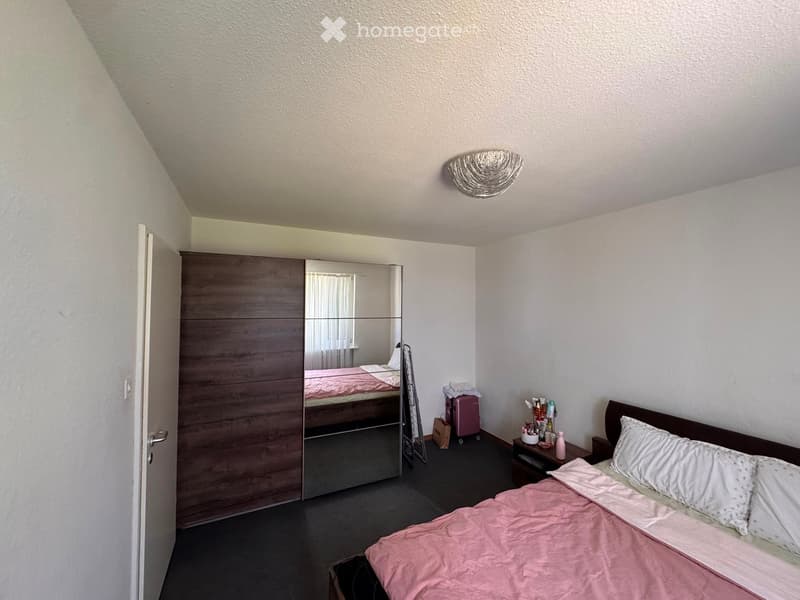 Bedroom with a double bed, pink bedding, a wardrobe with a mirror, a nightstand with a lamp, and a small table with personal items.