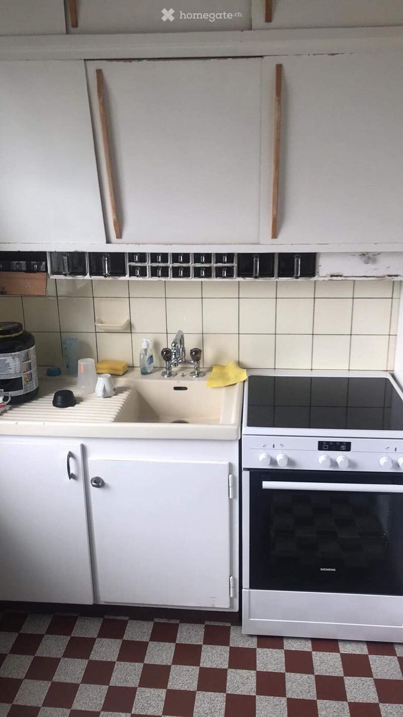 A small kitchen with white cabinets, a sink, a faucet, and an oven. The countertop has various items including a bottle, a cup, and a spoon.