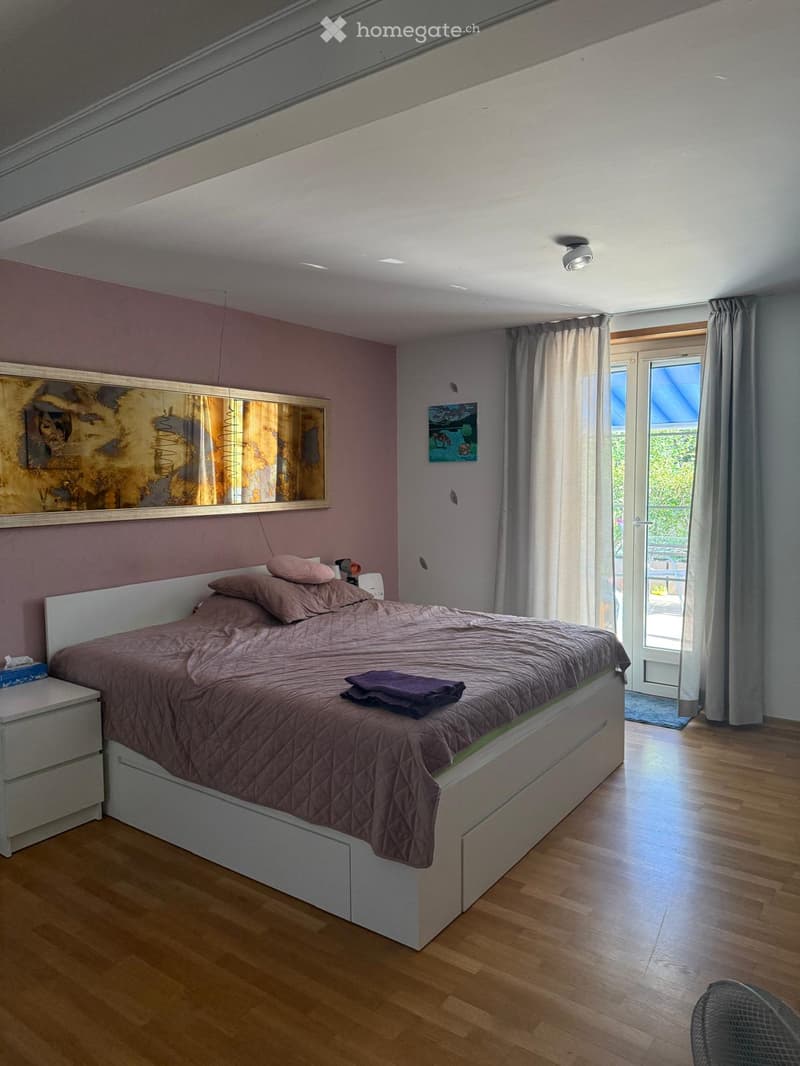 Modern bedroom with a large bed, nightstand, mirror, and artwork on the walls. The room features wooden flooring and large windows with curtains.