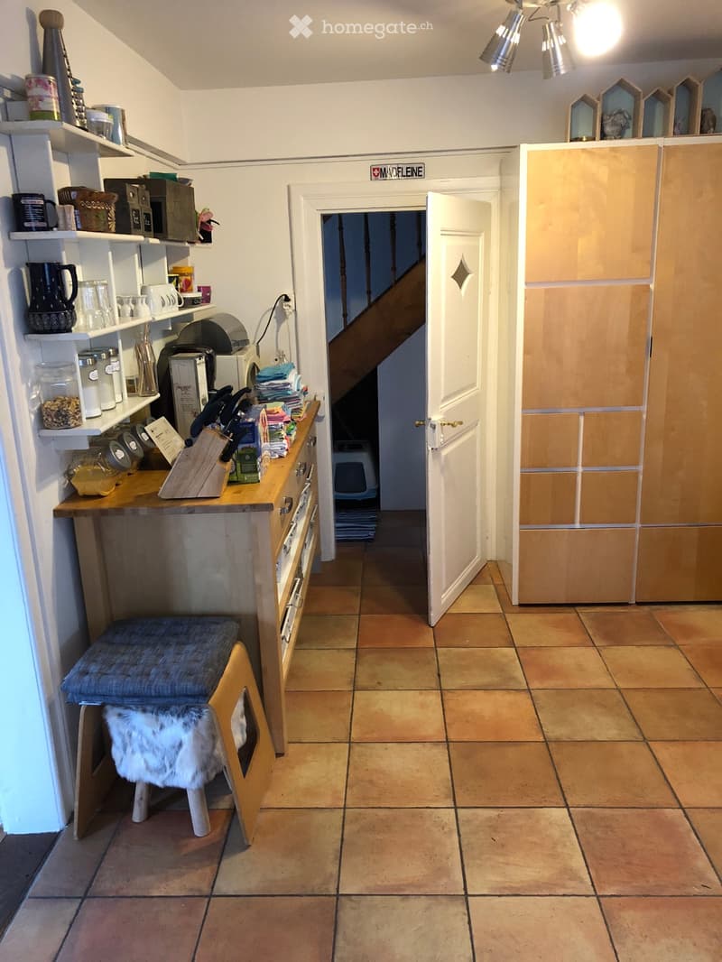 A hallway with tiled flooring, a small wooden bench with a fur throw, and a doorway leading to another room. Shelves with various items are visible on the left side.