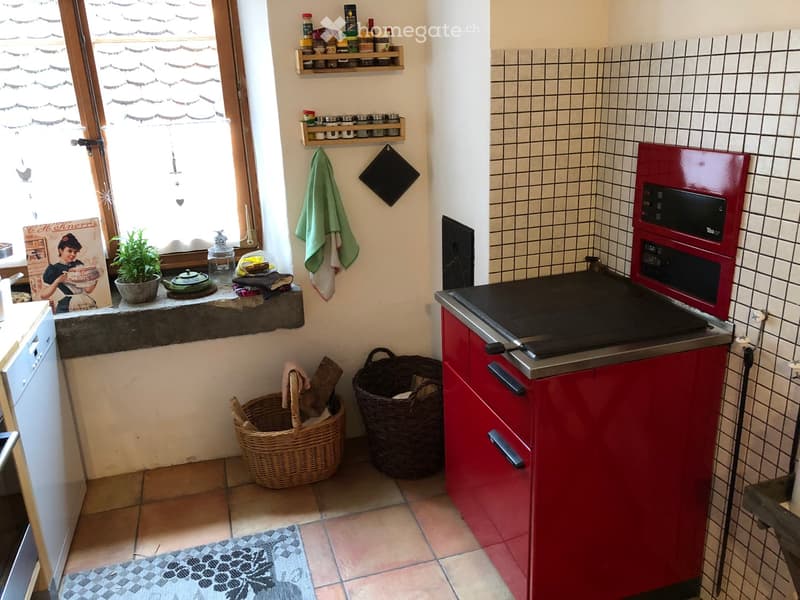 A cozy kitchen with red cabinets, a black countertop, and a tiled backsplash. Features include a built-in oven, a dishwasher, wicker baskets, a small plant on the windowsill, and various kitchen utensils and decorations.