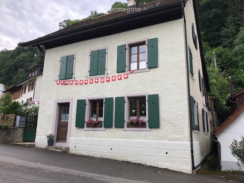 Two-story white house with green shutters, pink window frames, and flower boxes. Features a wooden front door, decorative Swiss flag bunting, and a small garden area.