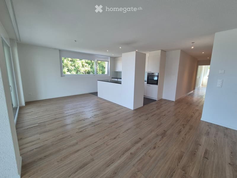Empty kitchen with wooden flooring, white cabinets, and a large window overlooking greenery.