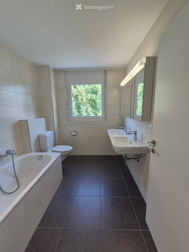 Modern bathroom with a bathtub, toilet, double sink, and large mirror. The space features tiled walls and dark tiled flooring, with a window providing natural light.