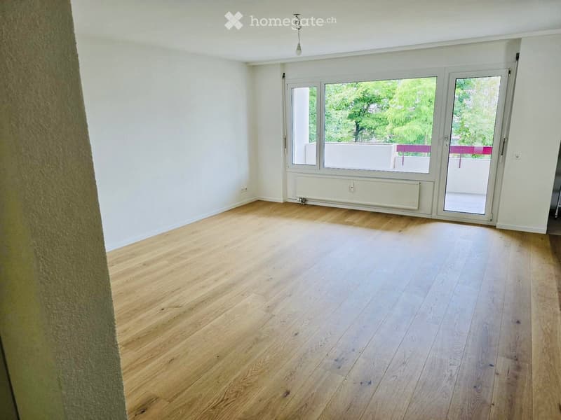 Empty room with light wooden flooring, white walls, large windows, and a sliding glass door leading to a balcony.