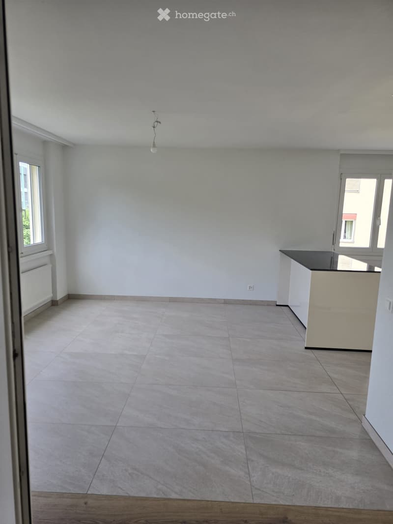Empty room with tiled floor, white walls, and a small kitchen counter in the corner. Windows on two walls allow natural light.