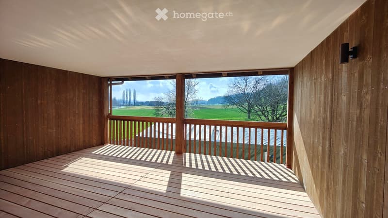 Wooden balcony with a railing, overlooking a green field and trees under a partly cloudy sky.