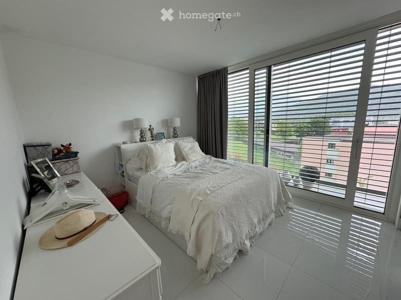 Modern bedroom with a large bed, nightstands, lamps, a small table with a hat, and a view through large windows.