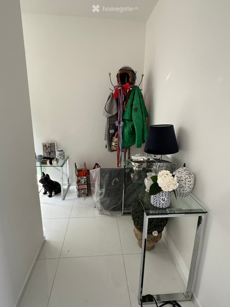 A modern hallway with white tiled flooring, a glass table with a lamp, vase, teapot, and picture frame, a coat rack with jackets, and a small dog sitting near a side table.