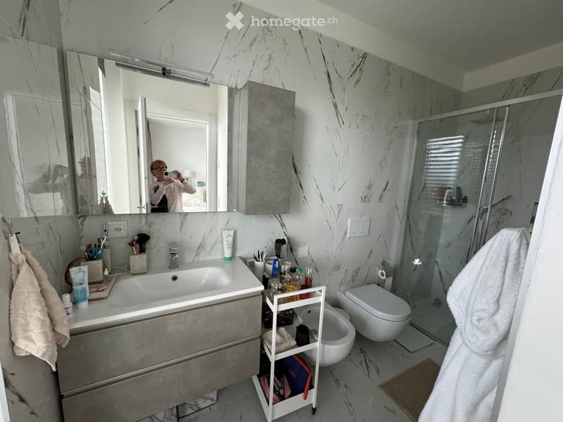 Modern bathroom with marble tiles, a large sink with a mirror, a toilet, a bidet, a shower stall, and a towel on the floor.