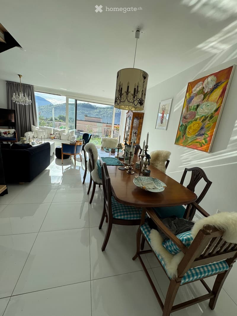 Bright dining area with a wooden dining table, chairs with fur cushions, a sofa, a chandelier, and a view through large windows.