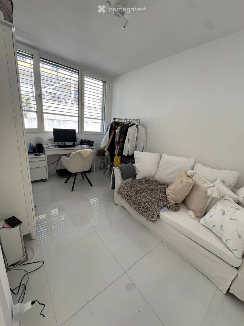A modern, multi-functional room with a sofa, desk, and clothing rack. The space features white walls, a glossy white tile floor, and large windows with blinds.