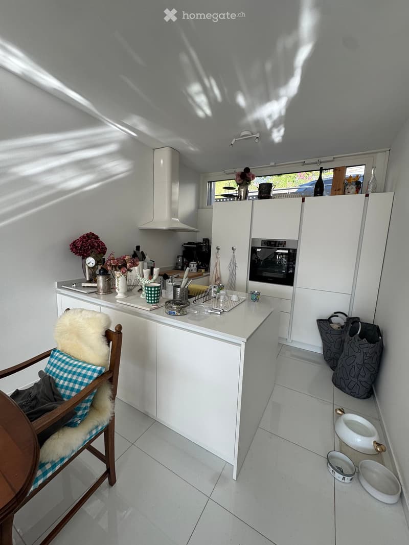 Bright kitchen with white cabinets, a central island, and various kitchen items on the counter. There is a chair with a fur throw, a handbag, and pet bowls on the floor.