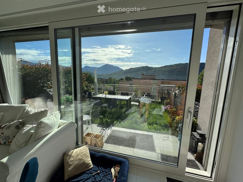 Bright living room with large windows offering a scenic mountain view, furnished with a sofa, chair, and a small table on the balcony.