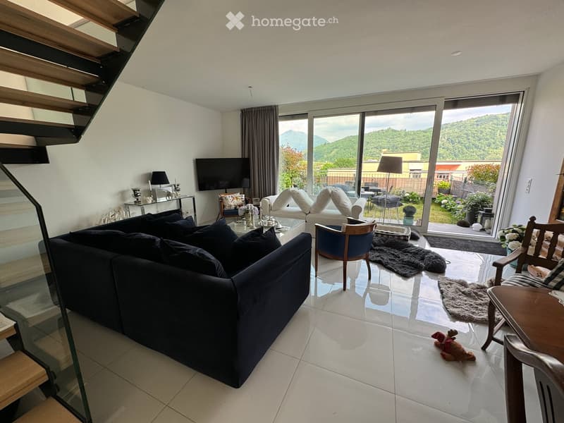Modern living room with large windows offering a scenic mountain view, featuring a dark blue sectional sofa, a wooden chair, a glass coffee table, a flat-screen TV, and decorative lamps.
