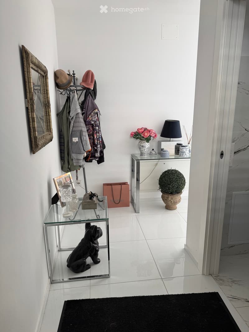 A modern hallway with white walls, a glass table, a coat rack, a mirror, a lamp, a vase with flowers, and a small statue.