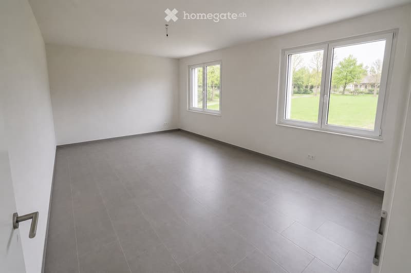 Empty room with grey tiled floor, white walls, and large windows overlooking a green area.