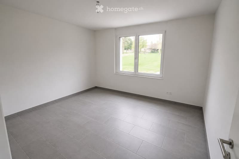 Empty room with white walls, grey tiled floor, and a large window showing a green outdoor view.