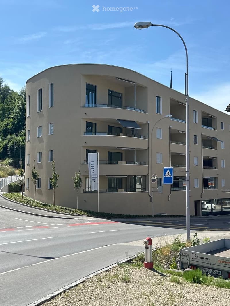 Modern multi-story residential building with balconies, large windows, and a curved facade. The building is surrounded by greenery and a paved street.