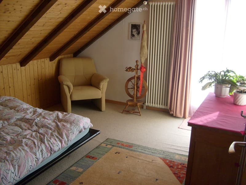 Cozy attic bedroom with wooden slanted ceilings, a bed with a floral duvet, a beige armchair, a small table with potted plants, a spinning wheel, and a radiator.