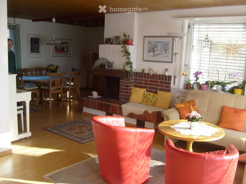 Bright living room with wooden floors, a brick fireplace, a cream sofa with orange cushions, a red armchair, a coffee table with a floral centerpiece, and a dining area with a table and chairs.