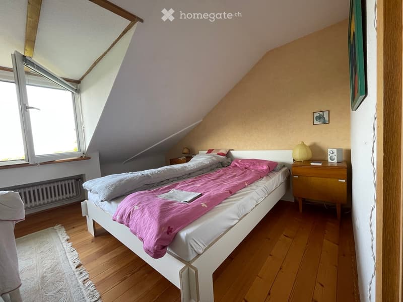 Cozy attic bedroom with wooden floors, slanted ceilings, and large windows. Features a double bed with pink bedding, a nightstand with a lamp and speaker, and a small picture frame on the wall.