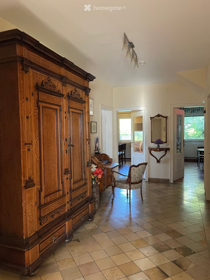Hallway with a large wooden cabinet, a patterned chair, a small table with a vase of flowers, and a mirror on the wall.