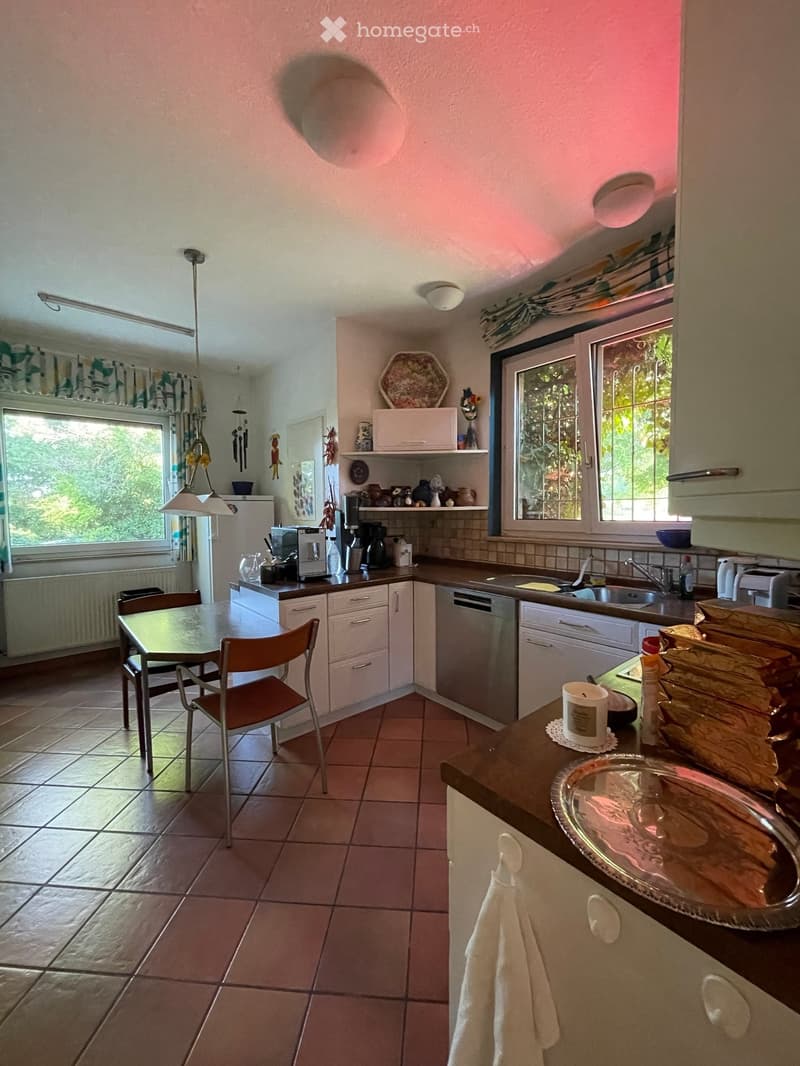 A well-lit kitchen with tiled floors, white cabinets, and various appliances including a dishwasher, refrigerator, microwave, and coffee maker. There is a dining area with a table and chairs, and decorative items on the countertops and shelves.
