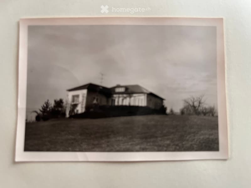 Black and white photograph of a two-story house with a pitched roof, visible windows, and a small porch area.