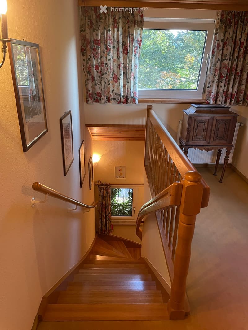 Wooden staircase with wooden handrails, floral curtains, framed pictures on the wall, and a small table with books in the background.