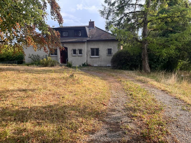 Two-story house with a gray exterior, multiple windows, and a small porch. The surrounding area includes a gravel path and grassy yard with trees.