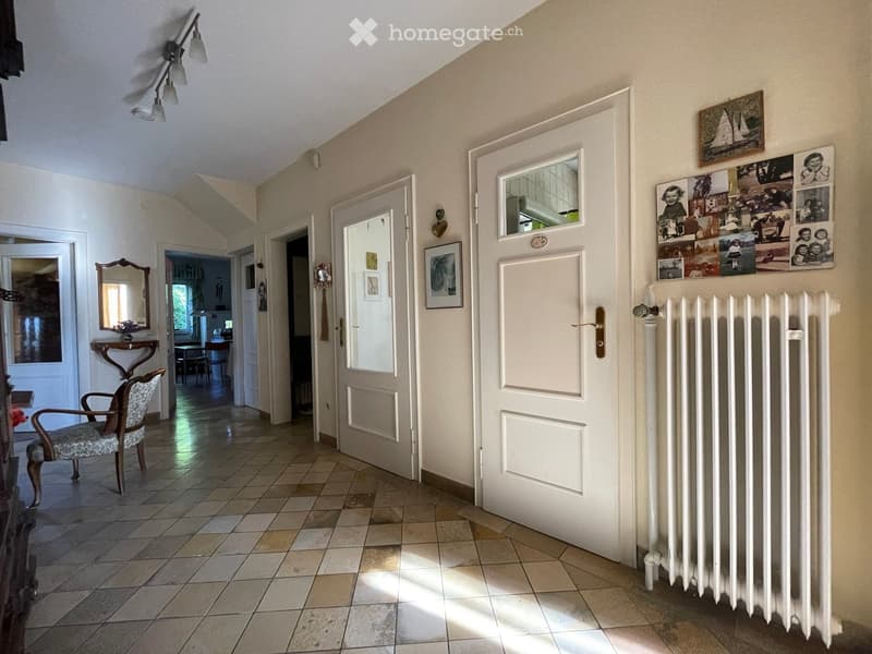 A well-lit hallway with tiled flooring, several doors leading to different rooms, a radiator on the right side, and a small table with a mirror and chair on the left side.