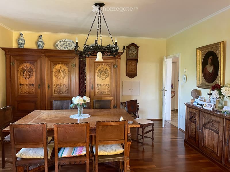 Elegant dining room with a wooden dining table surrounded by six chairs, a large wooden cabinet, a chandelier, and decorative vases on top of the cabinet. A framed portrait and a flower arrangement are visible on a sideboard.