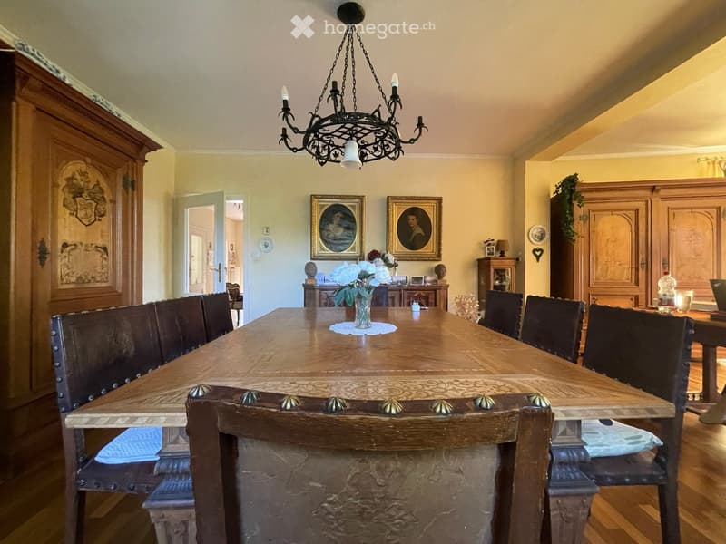 Elegant dining room with a long wooden dining table surrounded by dark wooden chairs, a chandelier hanging from the ceiling, and decorative cabinets and artwork on the walls.