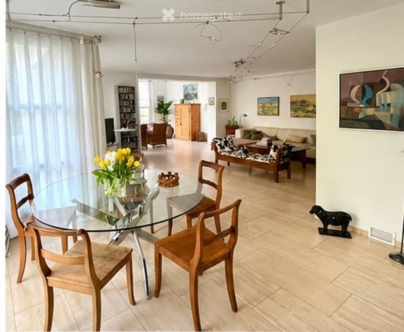 Bright, open-plan living and dining area with light wood flooring, large windows with white curtains, a glass dining table with wooden chairs, a sofa, coffee table, bookshelf, cabinet, and decorative items including a flower vase and picture frames.