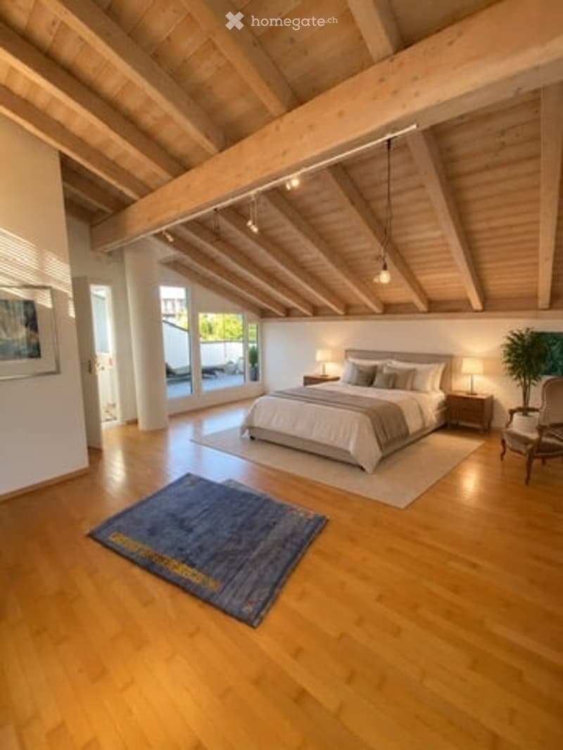Bright bedroom with wooden floors, exposed wooden beams, large windows offering a view, a neatly made bed with pillows and blankets, nightstands with lamps, a small chair, a blue rug, and a painting on the wall.