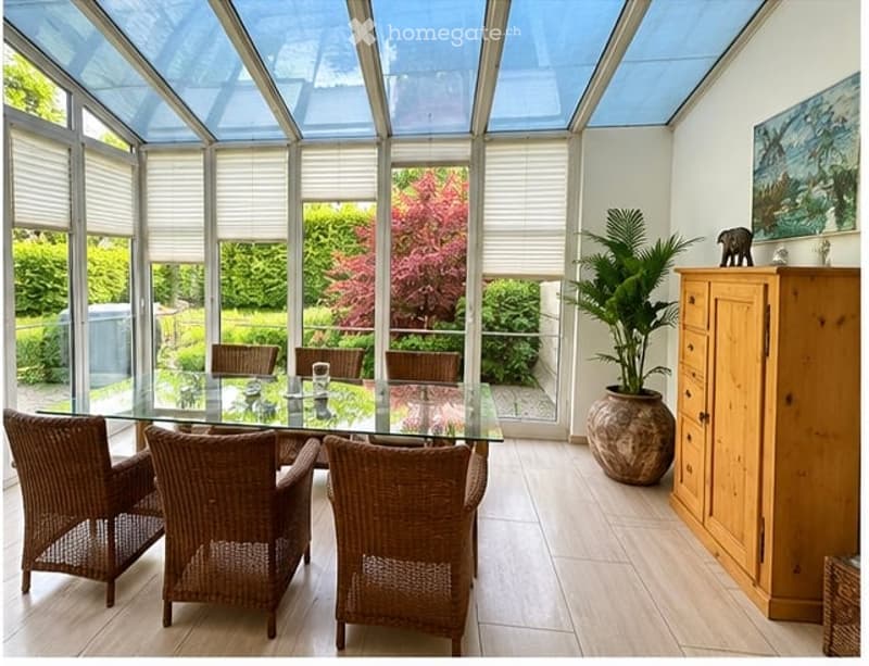 Bright sunroom with large windows and a glass ceiling, featuring a glass dining table surrounded by wicker chairs, a wooden cabinet, and a large potted plant.
