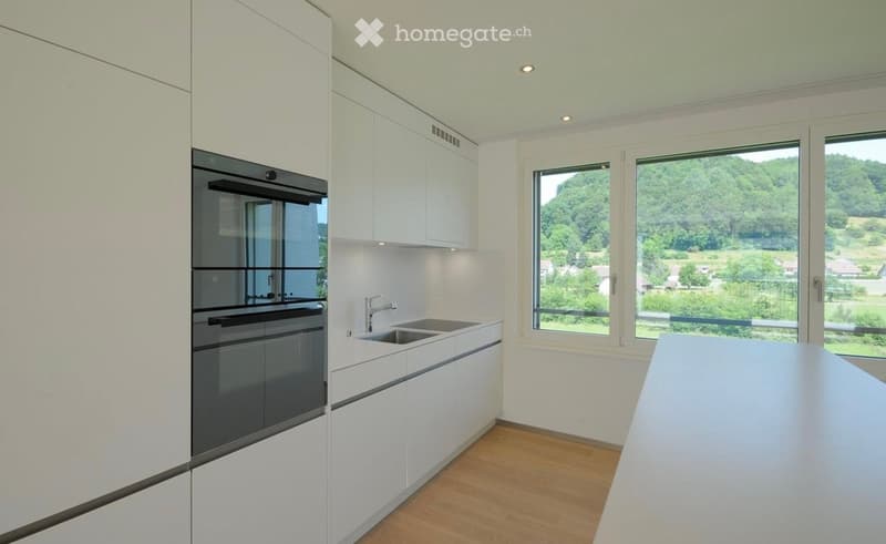 Modern kitchen with white cabinets, stainless steel oven, double sink, and large windows offering a scenic view of greenery and hills.