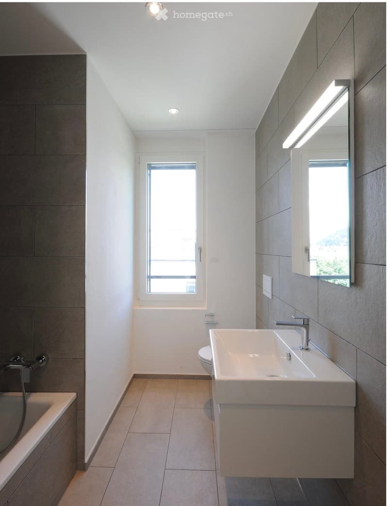 Modern bathroom with a white sink, bathtub, toilet, and large mirror. The space features light-colored tiles and ample natural light from a window.