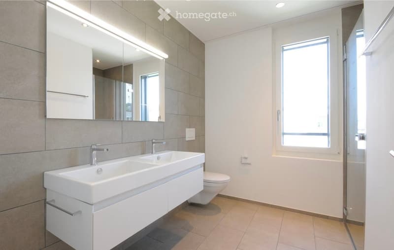 Modern bathroom with a double sink, large mirror, and a toilet. The space features light-colored tiles and a window providing natural light.