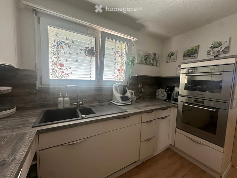 Modern kitchen with light-colored cabinets, a double sink, and stainless steel appliances including an oven and coffee maker. The countertops are made of wood, and there are decorative wall pieces above the cabinets.