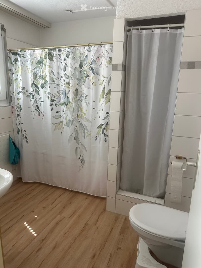 Bathroom with wooden flooring, white toilet, shower curtain with leaf design, and a sink visible on the left side.