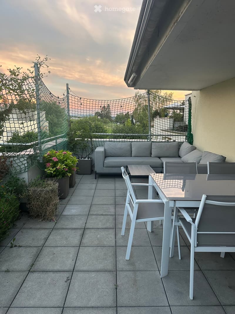 Balcony with grey sofa, small table, and white chairs, surrounded by potted plants and a net fence.