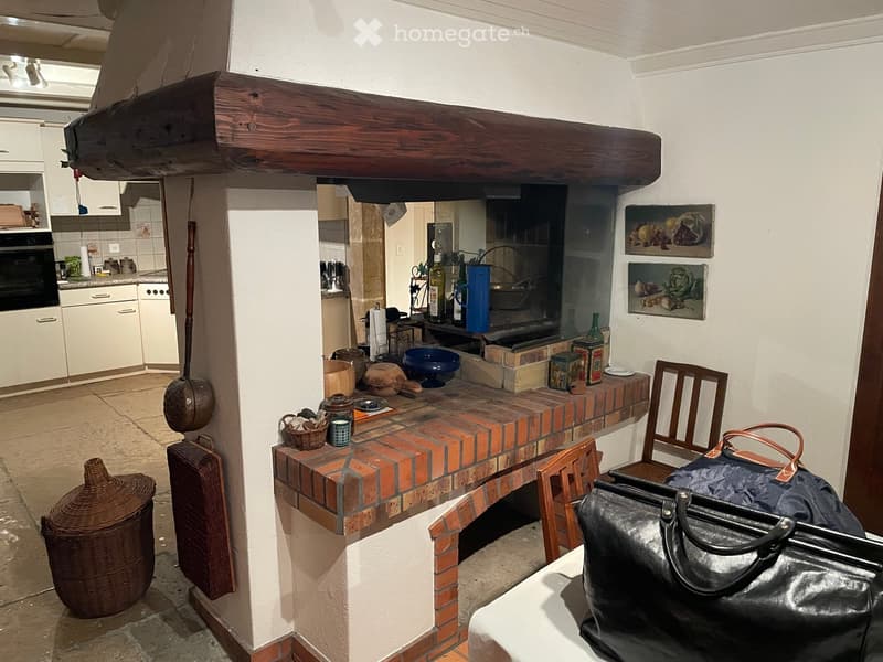 A rustic kitchen area with a brick countertop, wooden chairs, a handbag on a table, and various kitchen items including a pan, bottles, and a wicker basket.