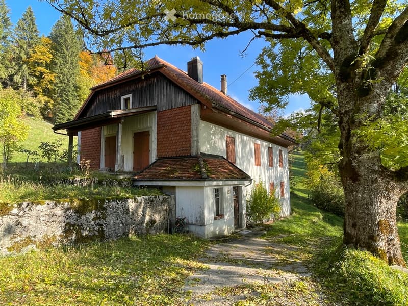 A two-story wooden house with a red-tiled roof, surrounded by trees and greenery in a rural setting.