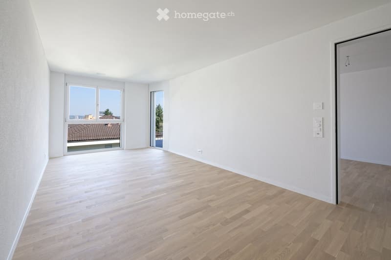Empty room with light wooden flooring and white walls, featuring large windows and a door leading outside.