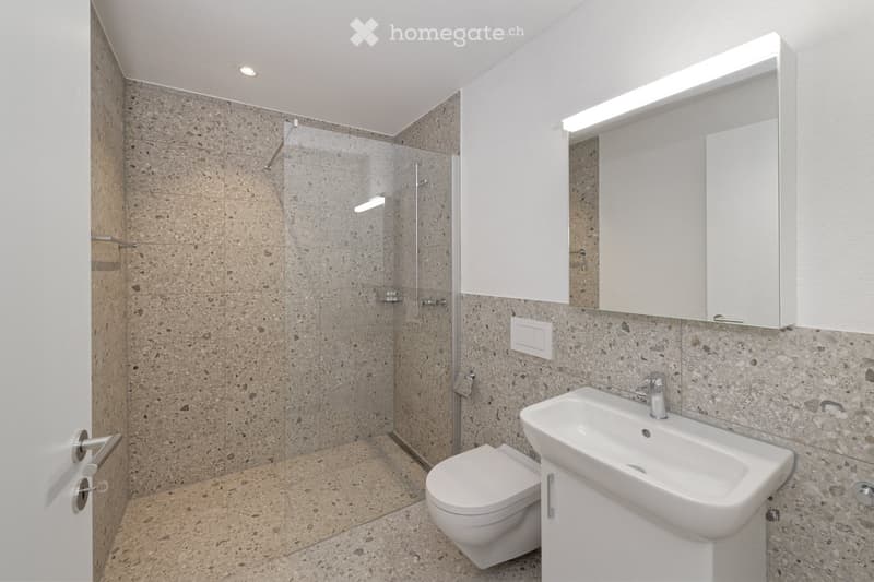 Modern bathroom with a white sink, toilet, and a glass-enclosed shower area. The walls and floor are tiled in a beige, speckled pattern, and a large mirror with built-in lighting is mounted above the sink.